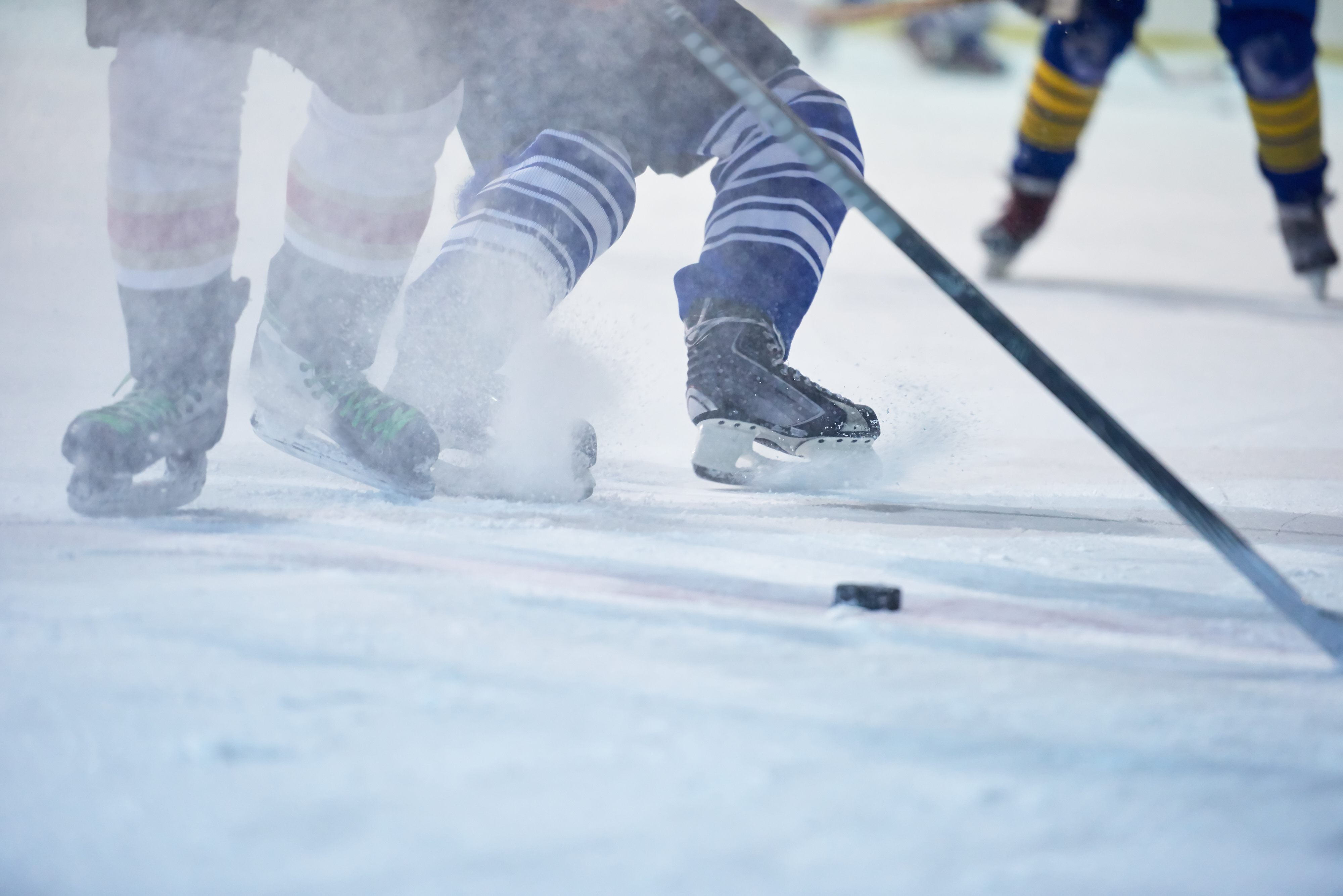 A hockey player's skates cutting into the ice and spraying snow, with the puck just ahead